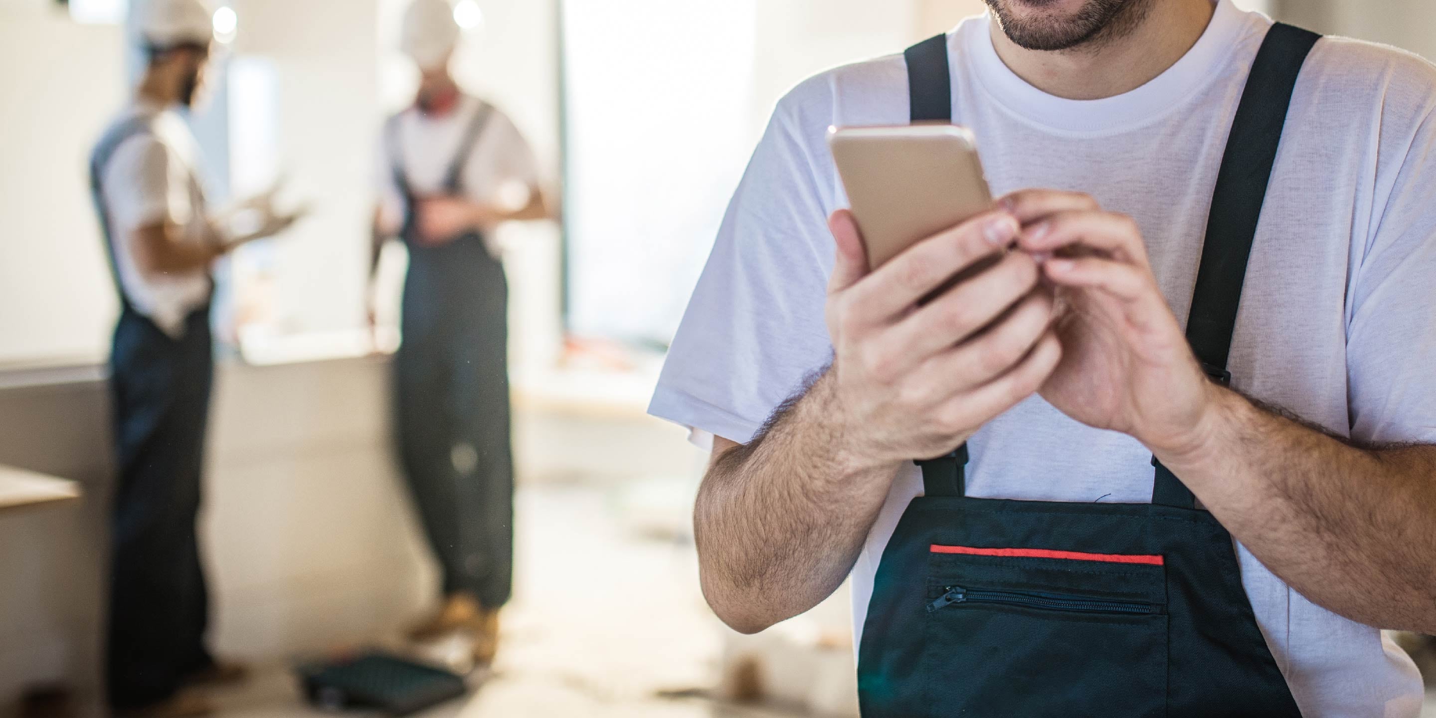 Happy male worker text messaging on smart phone while being in renovating home. There are people in the background.