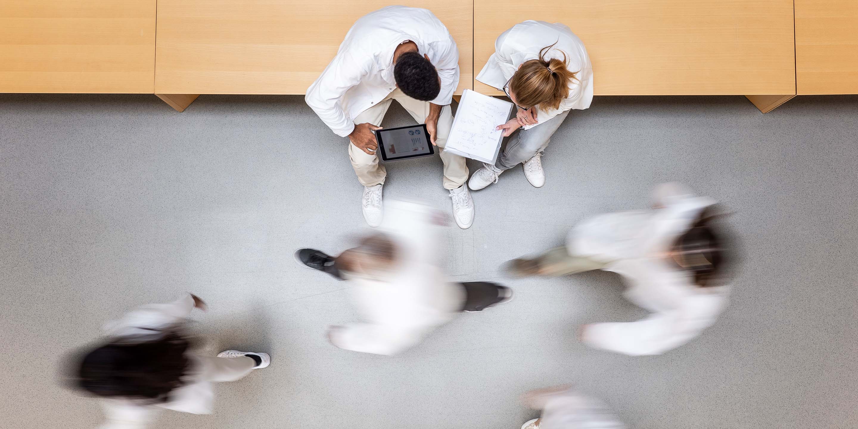 An overhead view of two medical professionals in white coats seated on a bench, one using a tablet and the other writing in a notebook. Four others walk by, creating a motion blur effect, in a bright indoor space with wooden benches and a light-colored floor.