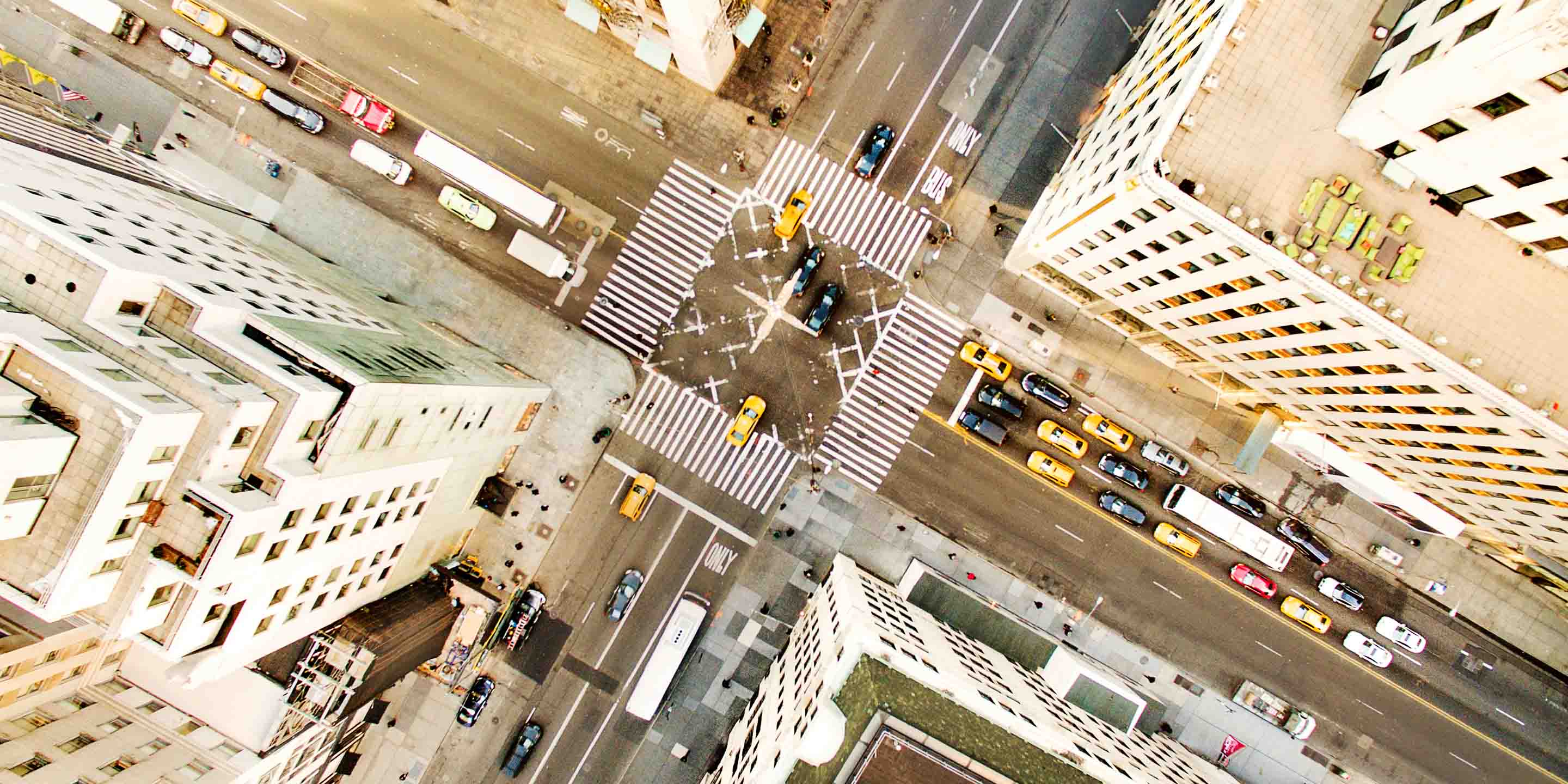 An overhead view of a bustling urban intersection featuring yellow taxis, cars, and buses navigating multiple lanes and crosswalks. Tall buildings surround the area, with rooftops and street markings clearly visible.