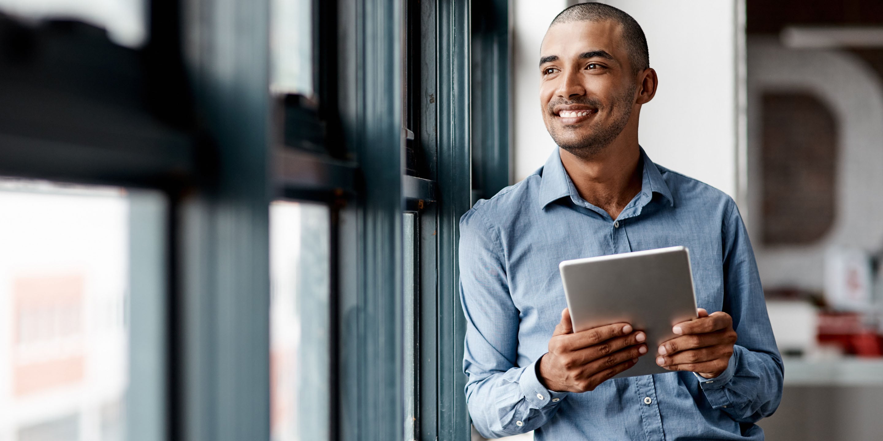 A person in a blue shirt stands by a window, holding and viewing a tablet. The face is blurred for privacy, and the setting suggests a modern, tech-savvy work environment with natural light.