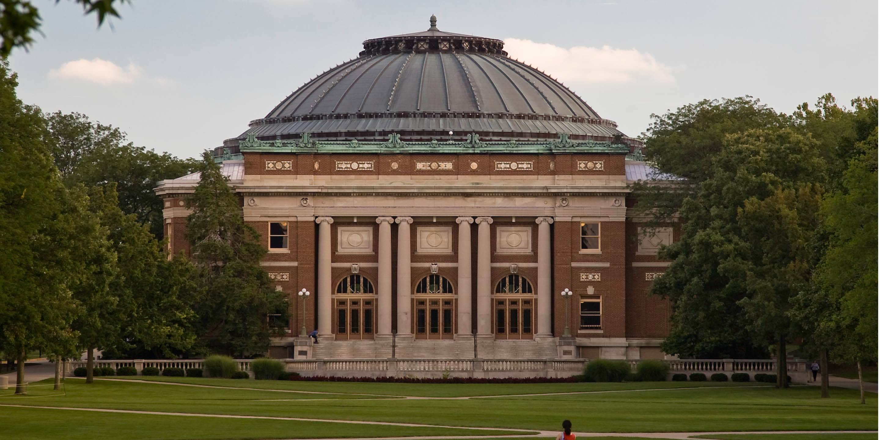 Brick building with dome, columns, trees, and green lawn.
