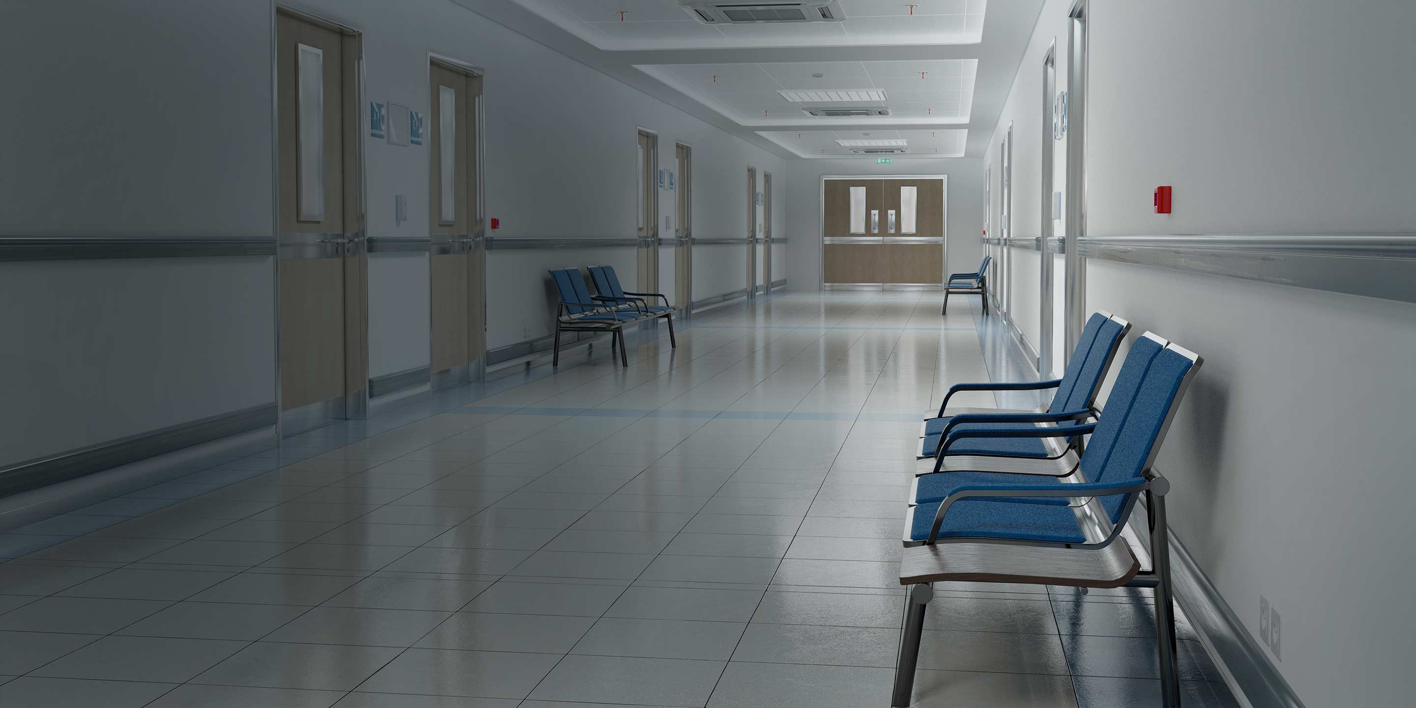 A young girl in an orange sweater is writing in a notebook. A long, white corridor with a few blue chairs against the wall and brown doors all the way at the end.