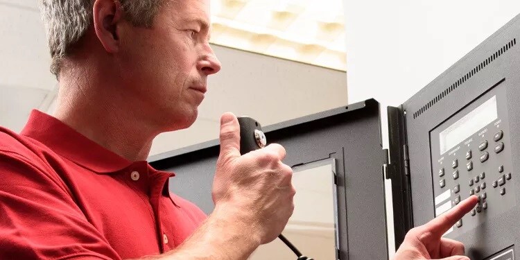 A person in a red shirt uses a microphone while pressing buttons on an electronic control panel. The face is blurred for privacy, and the setting suggests a technical or communication-focused environment.