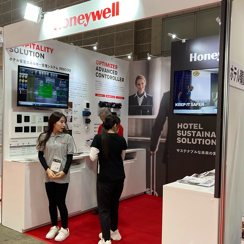 Photo of a Honeywell exhibition booth showcasing hospitality solutions, including the Optimizer Advanced Controller and Hotel Sustainability Solution. Features product displays, digital screens, and attendee interaction.