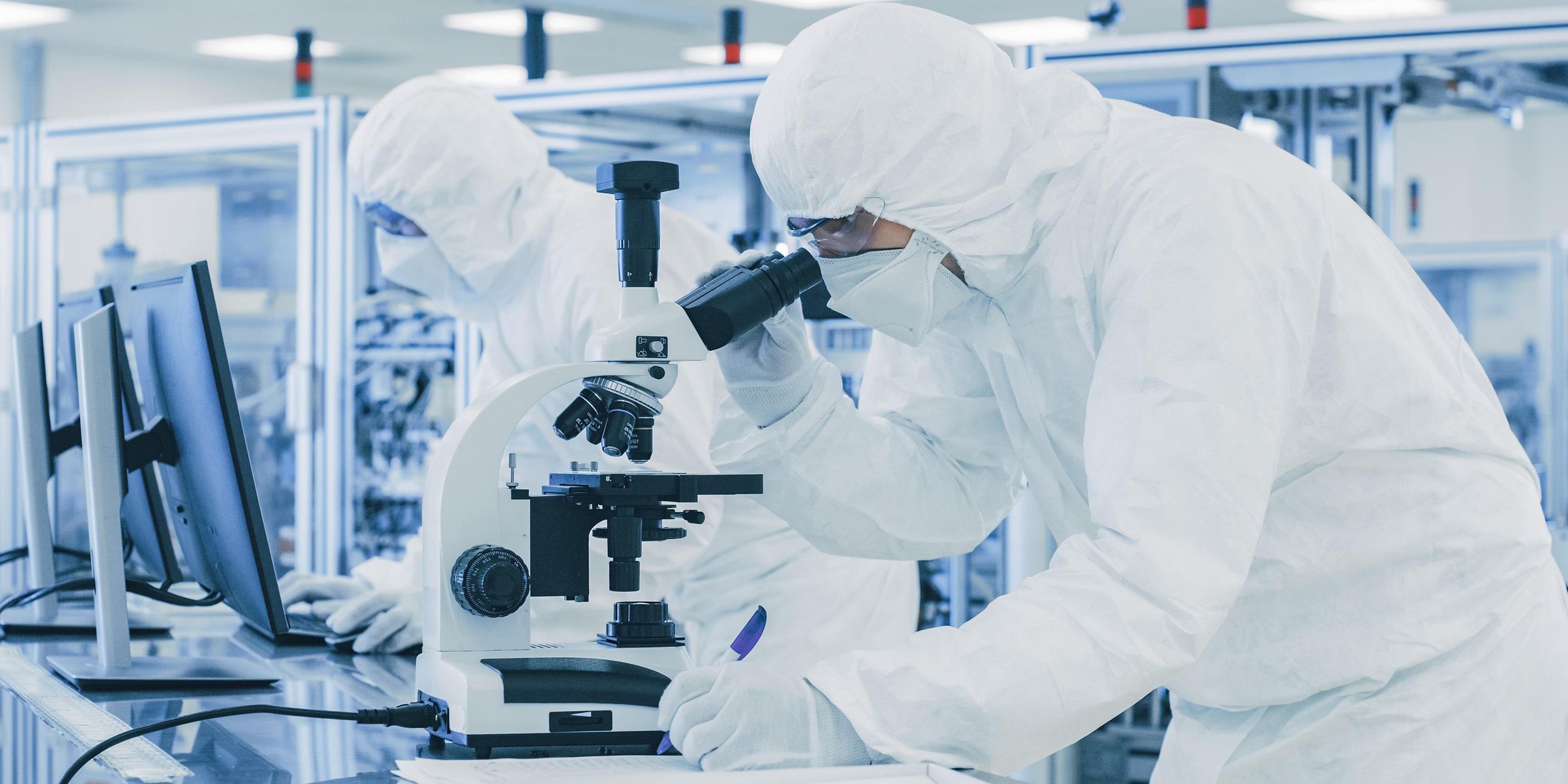 In Laboratory Scientist in Protective Clothes Doing Research, Using Microscope and Writing Down Data. Workers Working on a Modern Manufactory Producing Semiconductors and Pharmaceutical Items