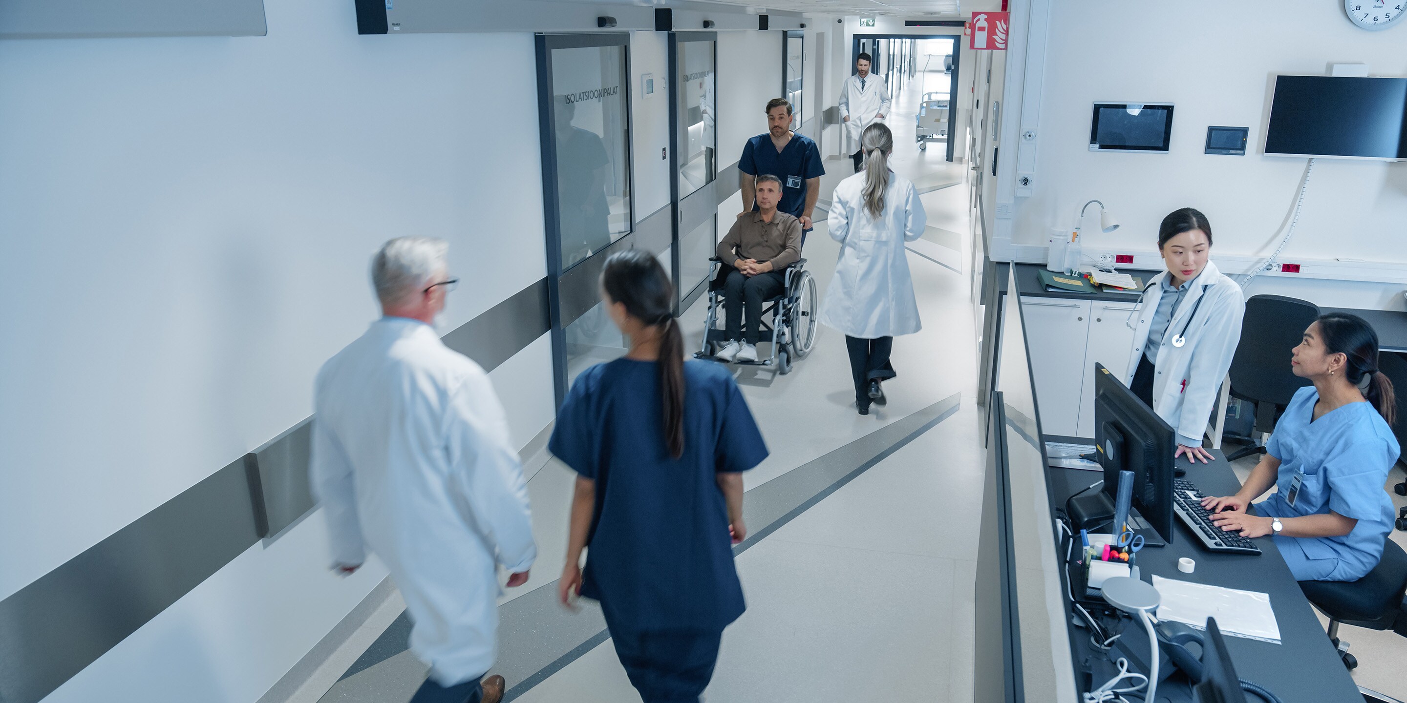 A busy hospital corridor featuring medical staff in white coats and scrubs attending to patients. A patient in a wheelchair is being escorted, with additional healthcare professionals walking and others at a reception desk. The environment portrays a professional healthcare setting.